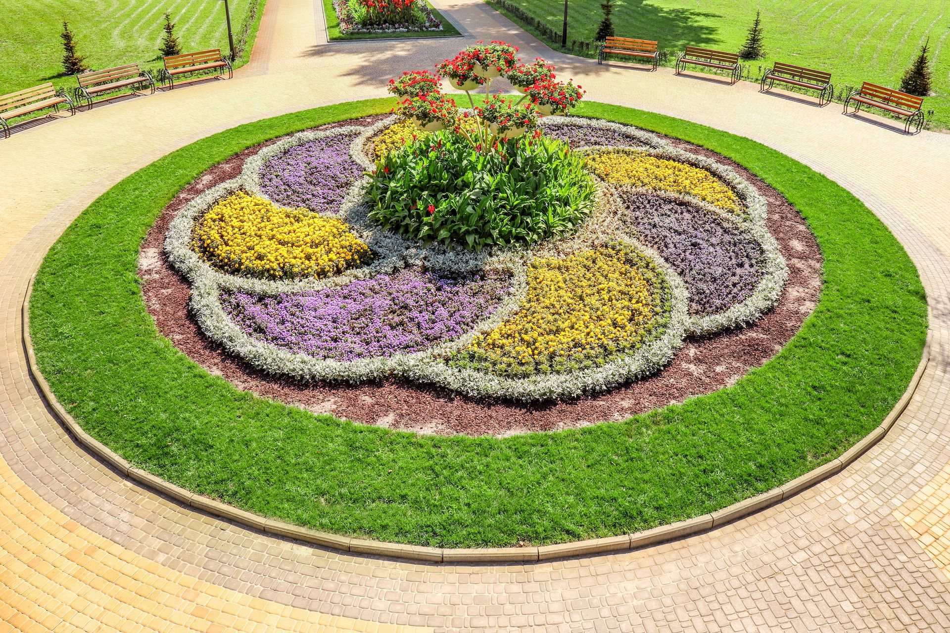 Round flower bed of yellow and purple annuals ringed by lawn and a paver plaza lined with benches