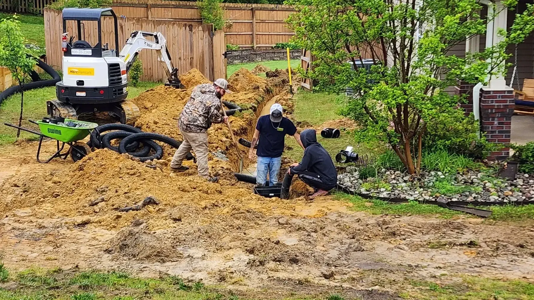 Construction workers installing drainage pipes near a fence with a small excavator on a muddy ground.