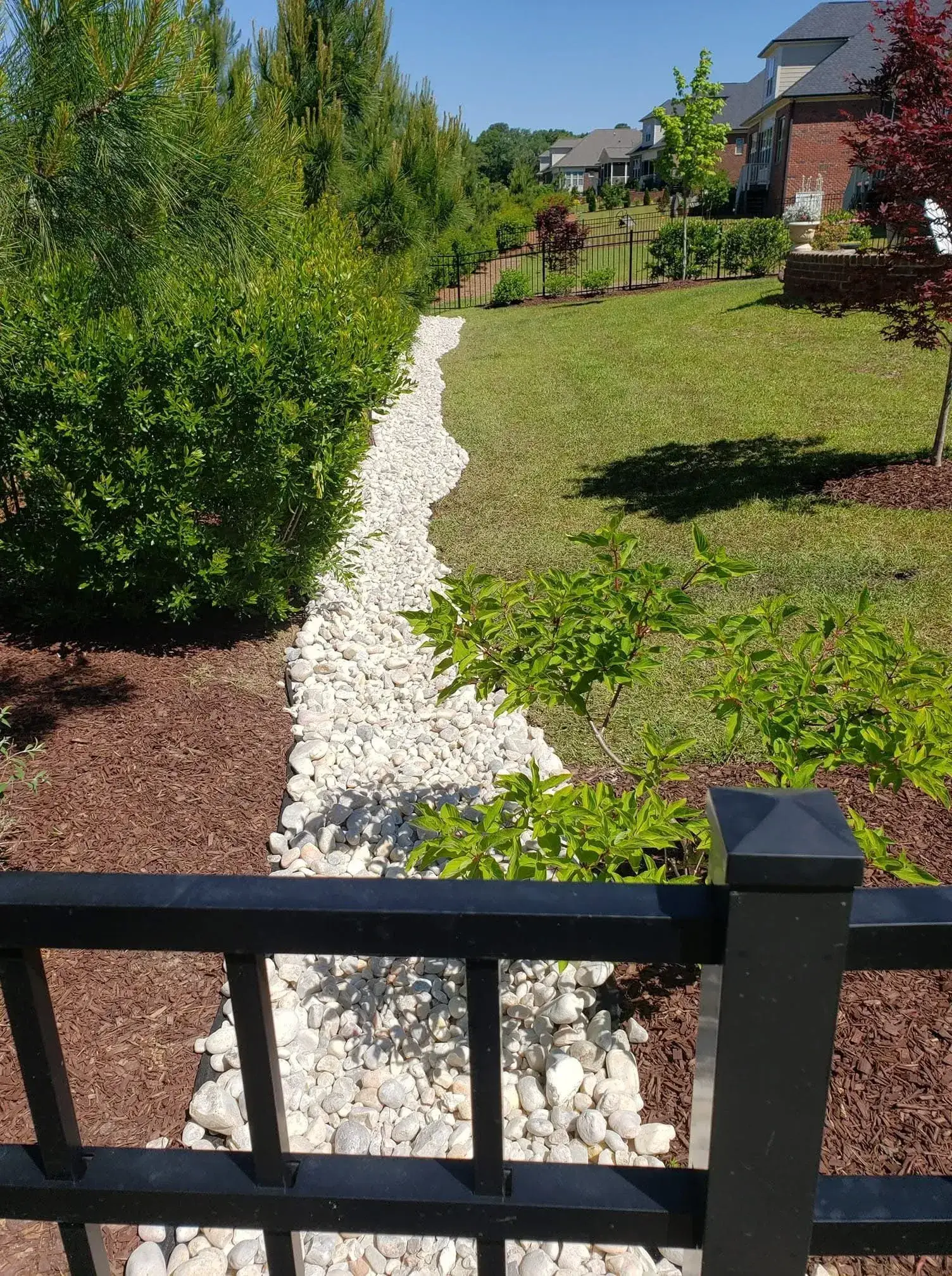 A backyard drainage channel lined with rocks, bordered by shrubs, mulch, and a black fence.
