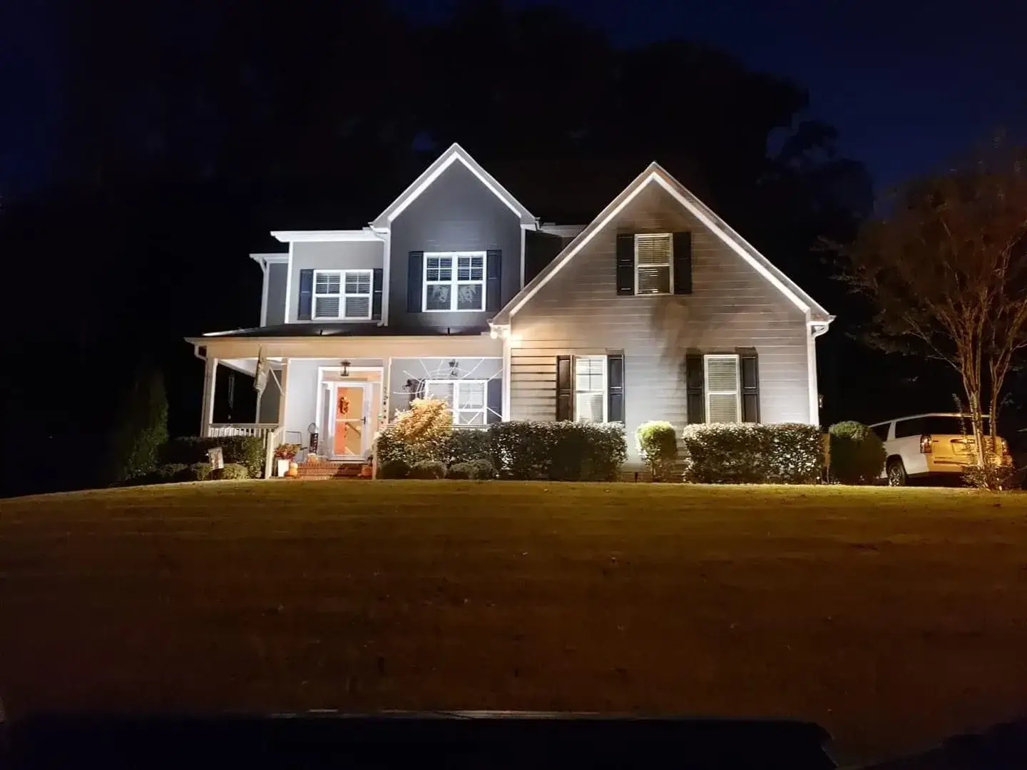 Two-story house illuminated at night with a front porch, bushes, and a vehicle on the side.