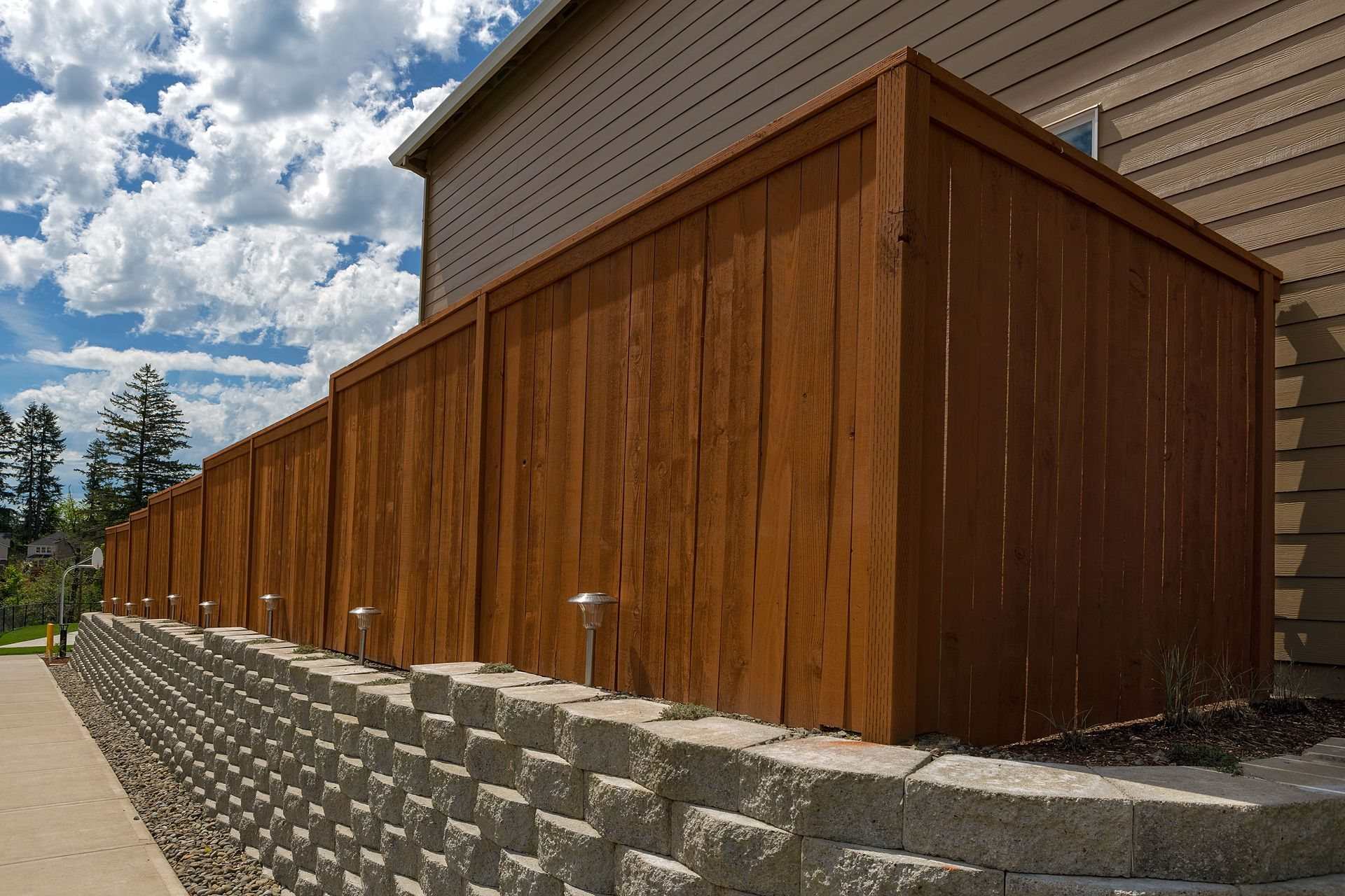 Tall block retaining wall with solar path lights below a stained wood privacy fence beside a house