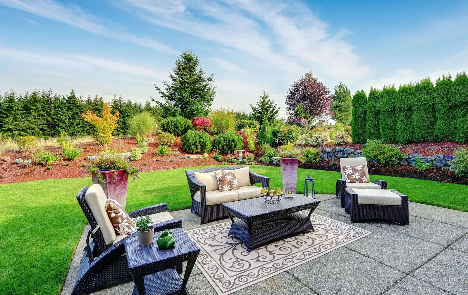 Patio with wicker furniture on stone, area rug, lawn, and landscaped garden under a blue sky.